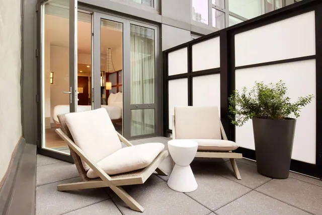 A private balcony with two cream lounge chairs, a white side table and a potted plant beside a black and white privacy screen, with glass doors open to a bedroom.