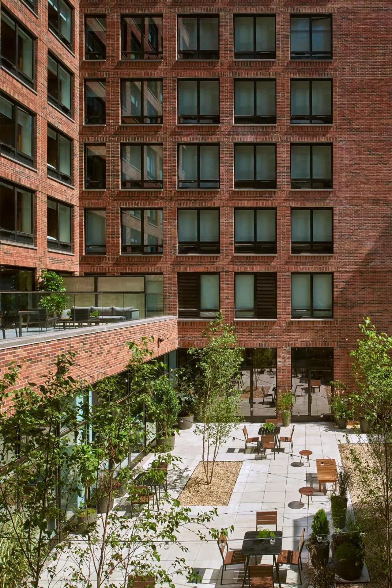 A multi-storey brick building with rows of windows overlooks a paved courtyard with outdoor tables, chairs and trees.