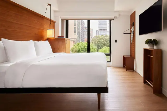 A bedroom with a made king bed, a wood headboard, a wall-mounted television and a large window looking onto city buildings and trees.