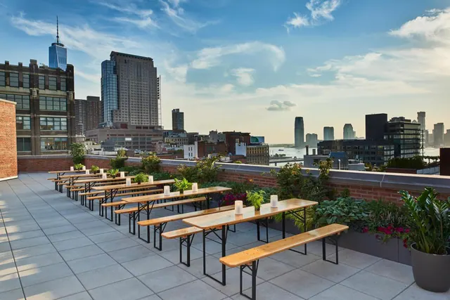 A rooftop terrace with long wooden communal tables, benches, candles and planters, overlooking a city skyline with One World Trade Center and a river.