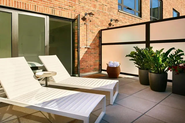 A paved terrace with two white sun loungers, a small round side table, potted plants, a basket of towels and a frosted glass privacy screen beside a brick wall.
