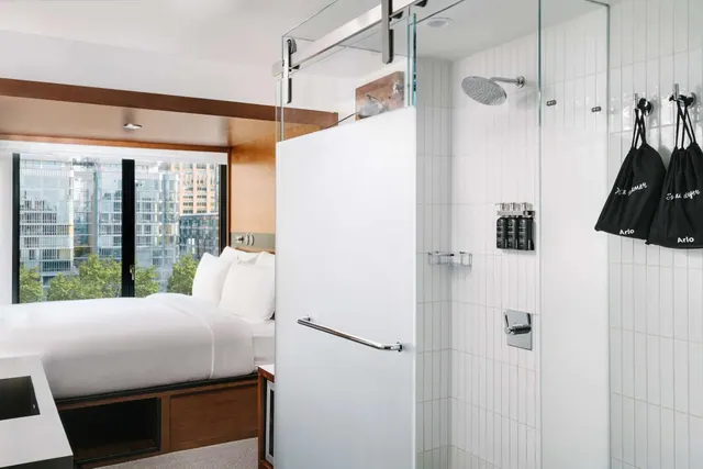 A made bed with white linens sits beside a large window looking onto city buildings and trees, with a glass-walled walk-in shower and white wall tiles beyond.
