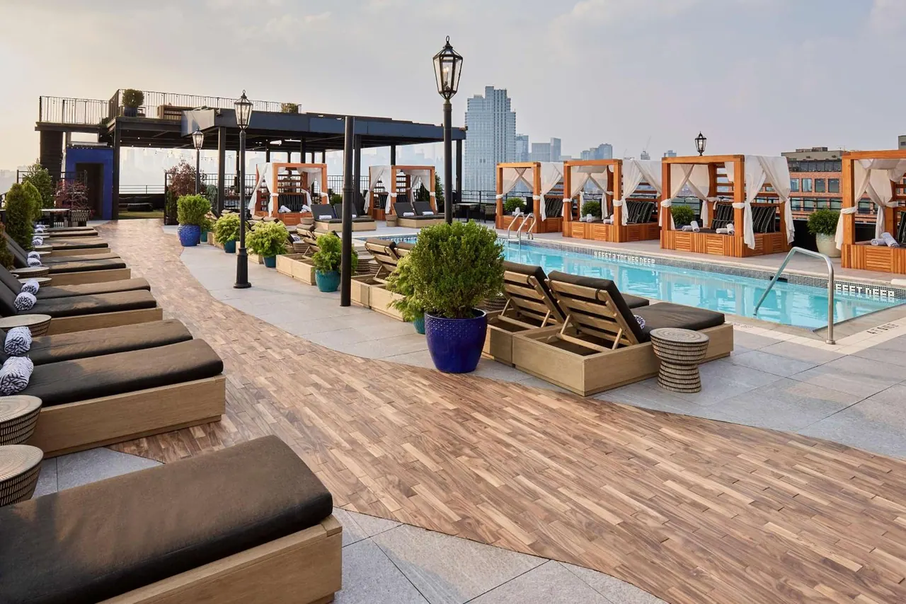 A rooftop pool deck with sun loungers, white-curtained cabanas, lamp posts and a city skyline in the hazy background.