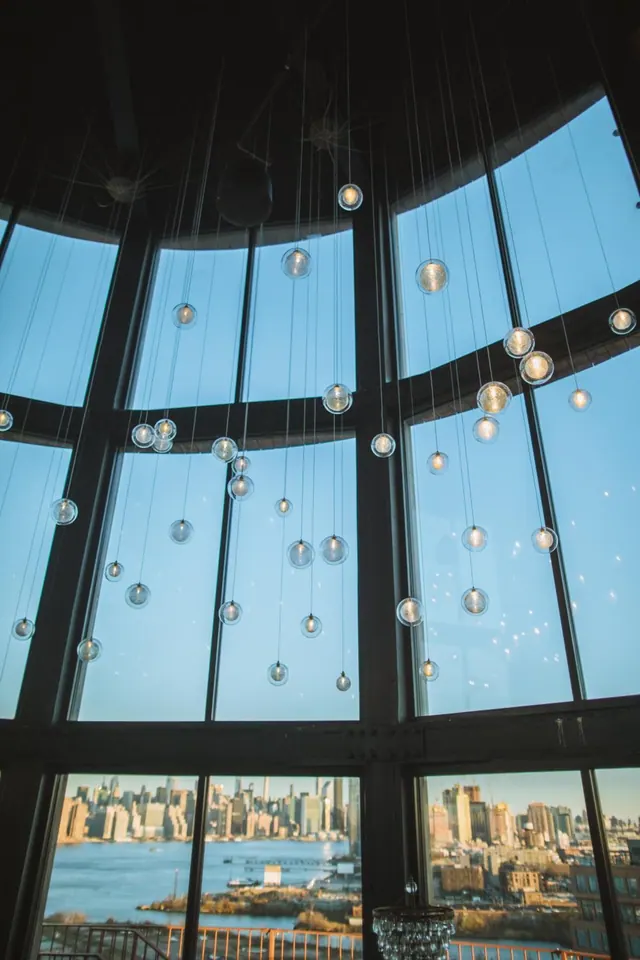 A round room with floor-to-ceiling curved windows and hanging glass globe lights, looking out over water toward a city skyline.