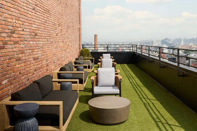 A rooftop terrace with artificial turf, dark sofas, armchairs and a round concrete coffee table, with a city skyline and a brick chimney beyond the railing.