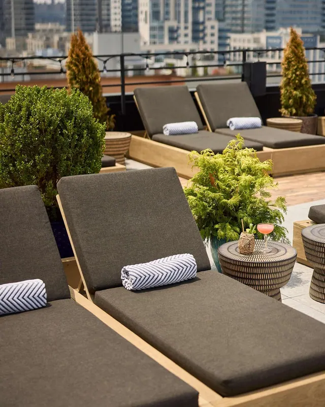 Rooftop terrace with grey-cushioned sun loungers, rolled striped towels, potted plants and side tables, with a city skyline of tall buildings in the background.