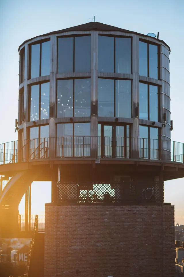 A cylindrical glass-walled tower with a railed balcony sits on a brick base, lit by low sunlight against a pale sky.