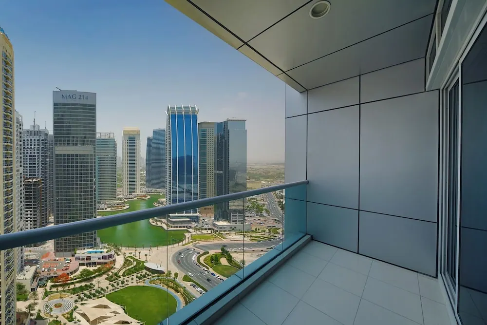 A glass-railed balcony with a tiled floor overlooks high-rise towers, a green lake and a landscaped park.