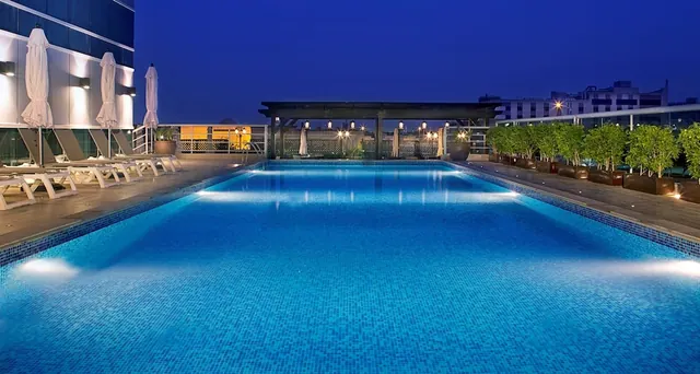 A rooftop swimming pool lit from below at dusk, with sun loungers, closed umbrellas, a covered pergola and potted trees beside it, with city buildings in the background.