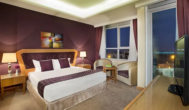 A bedroom with a king bed, two armchairs by a window, a wall-mounted television and a view of city buildings at dusk.