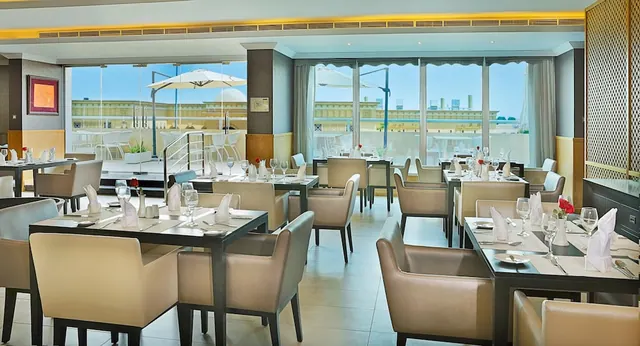 A restaurant dining room with set tables, leather armchairs and floor-to-ceiling windows overlooking an outdoor terrace and nearby buildings.