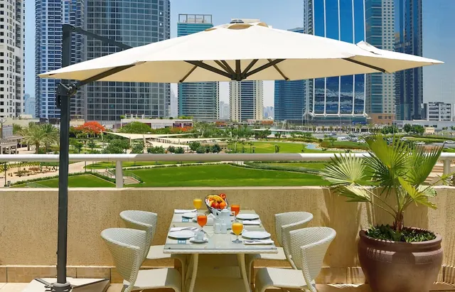 A breakfast table set with juice glasses, a fruit bowl and white wicker chairs sits on a balcony under a cantilever umbrella, overlooking a green park and high-rise towers.