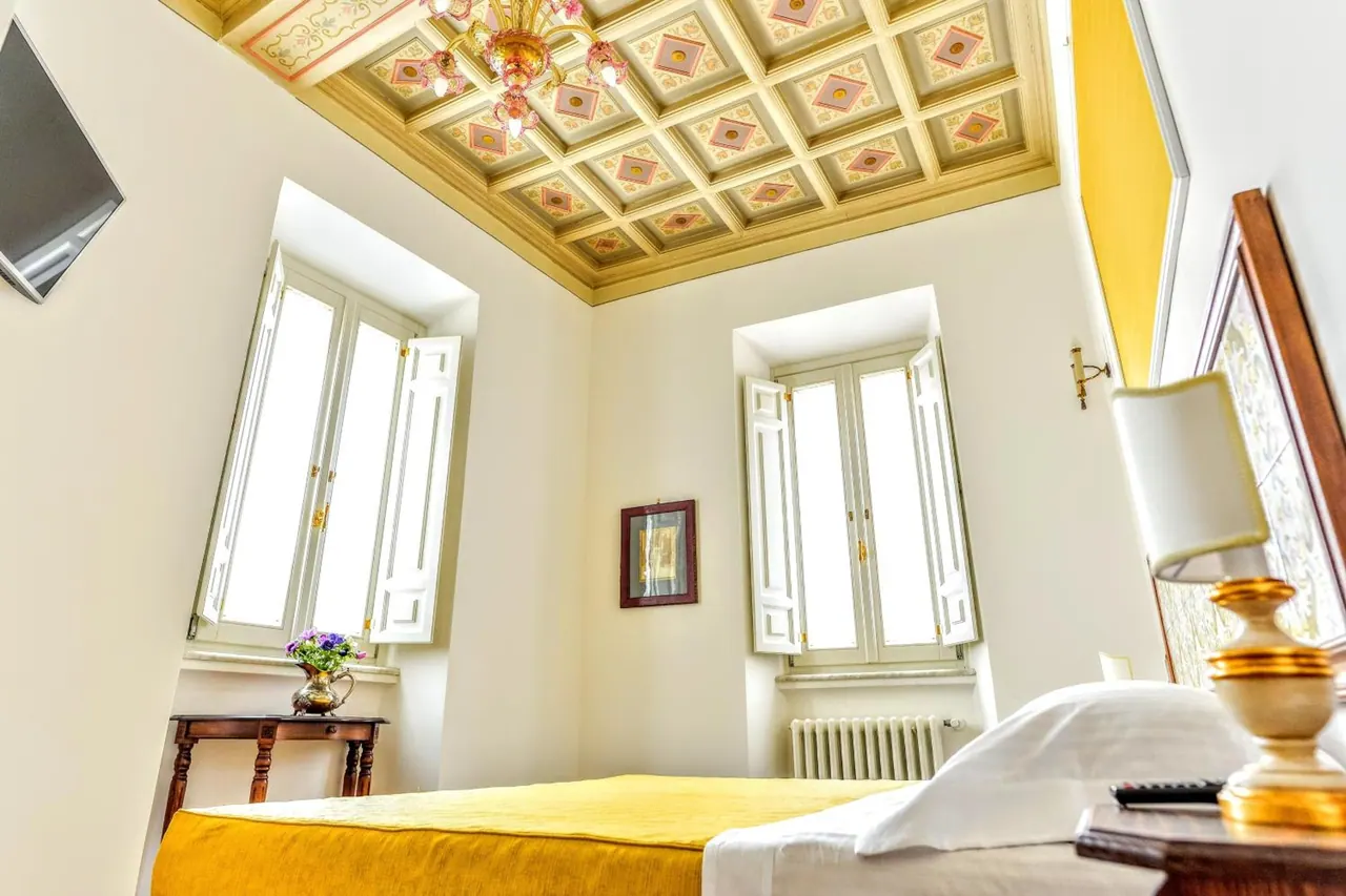 A bedroom with a yellow bedspread, a painted coffered ceiling with a chandelier, and two open shuttered windows.