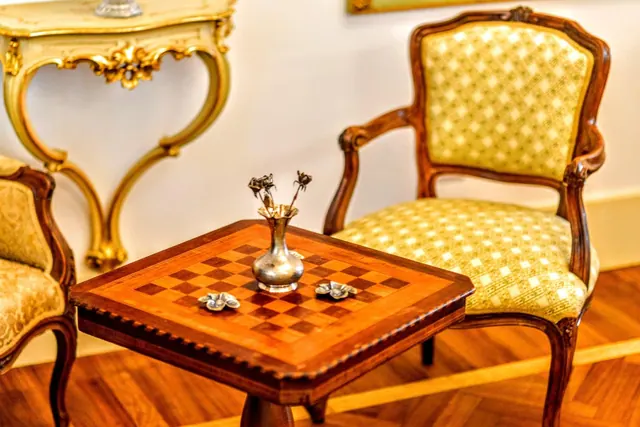 A chessboard-top side table with a silver vase sits beside an upholstered armchair with a carved wooden frame and a gilded console table against a cream wall.