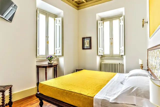 A bedroom with a yellow bedspread on a double bed, two open windows with white shutters, a radiator, and a wall-mounted television.