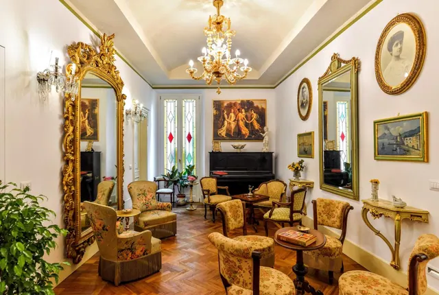 A formal salon with gilded mirrors, a crystal chandelier, upholstered armchairs, a black piano and a painting of dancing figures on the back wall.