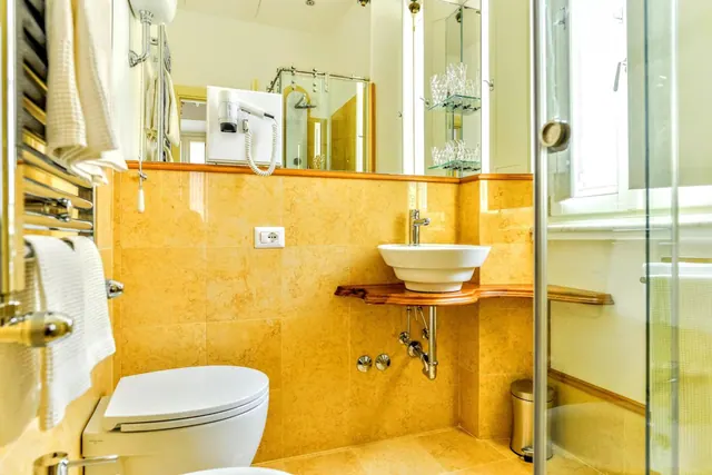 A bathroom with yellow stone-effect tiles, a wall-mounted toilet, a vessel sink on a wooden shelf, a wall hair dryer, a heated towel rail and a glass-walled shower.
