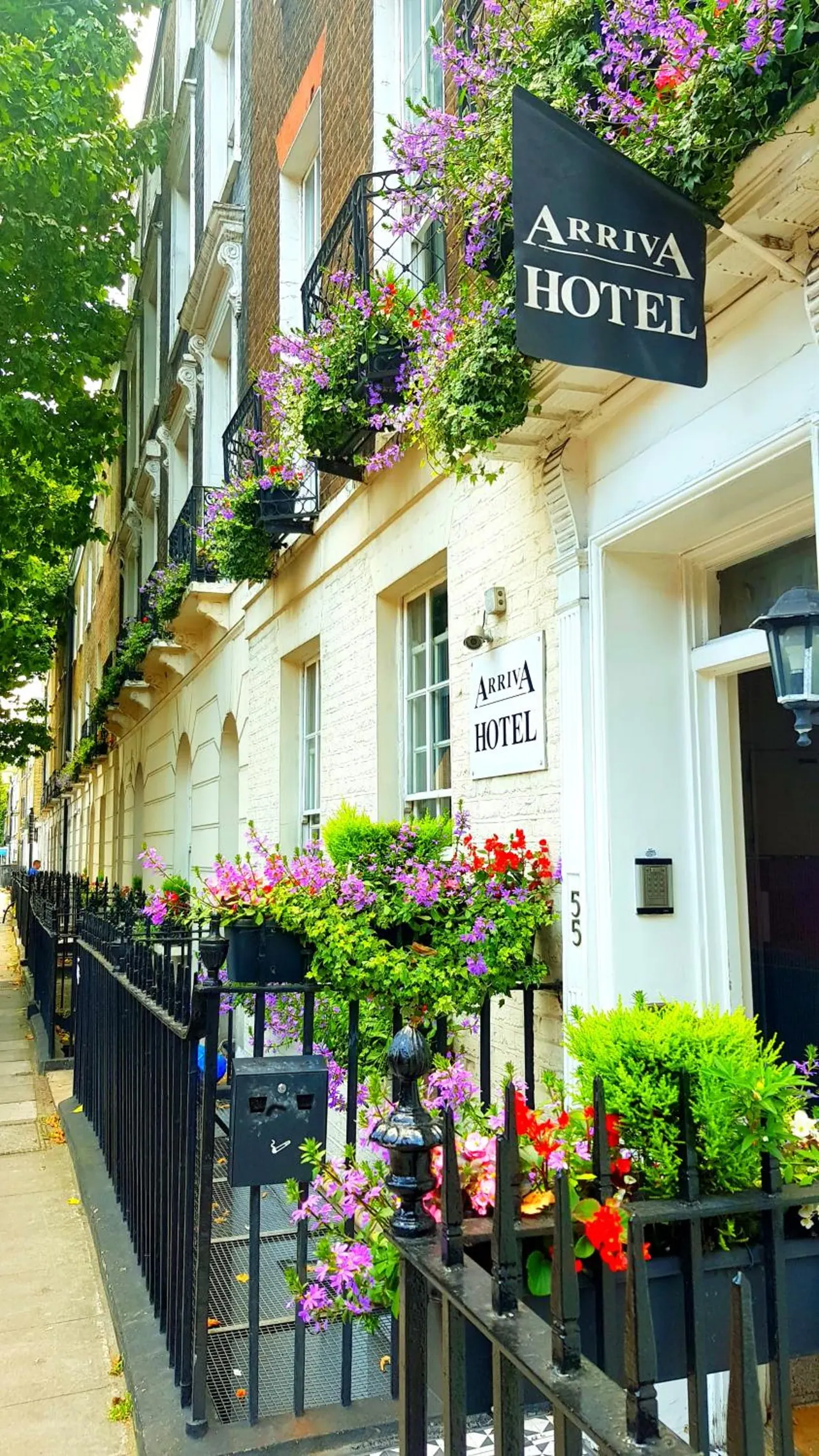 Street-facing front of the Arriva Hotel, with a black hanging sign, flower-filled window boxes and balconies, and black iron railings along the pavement.