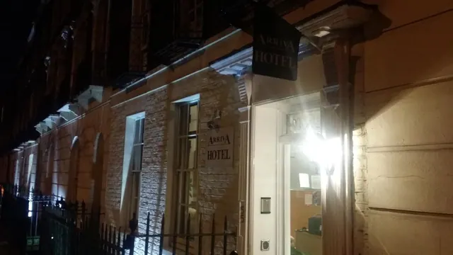 Night view of the Arriva Hotel facade with a hanging sign, a wall sign, an open entrance door and iron railings in front.