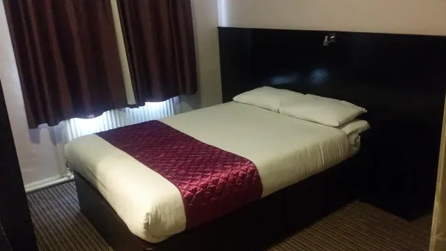 A double bed with cream bedding and a burgundy quilted runner sits beside brown curtains and a radiator in a dimly lit room.
