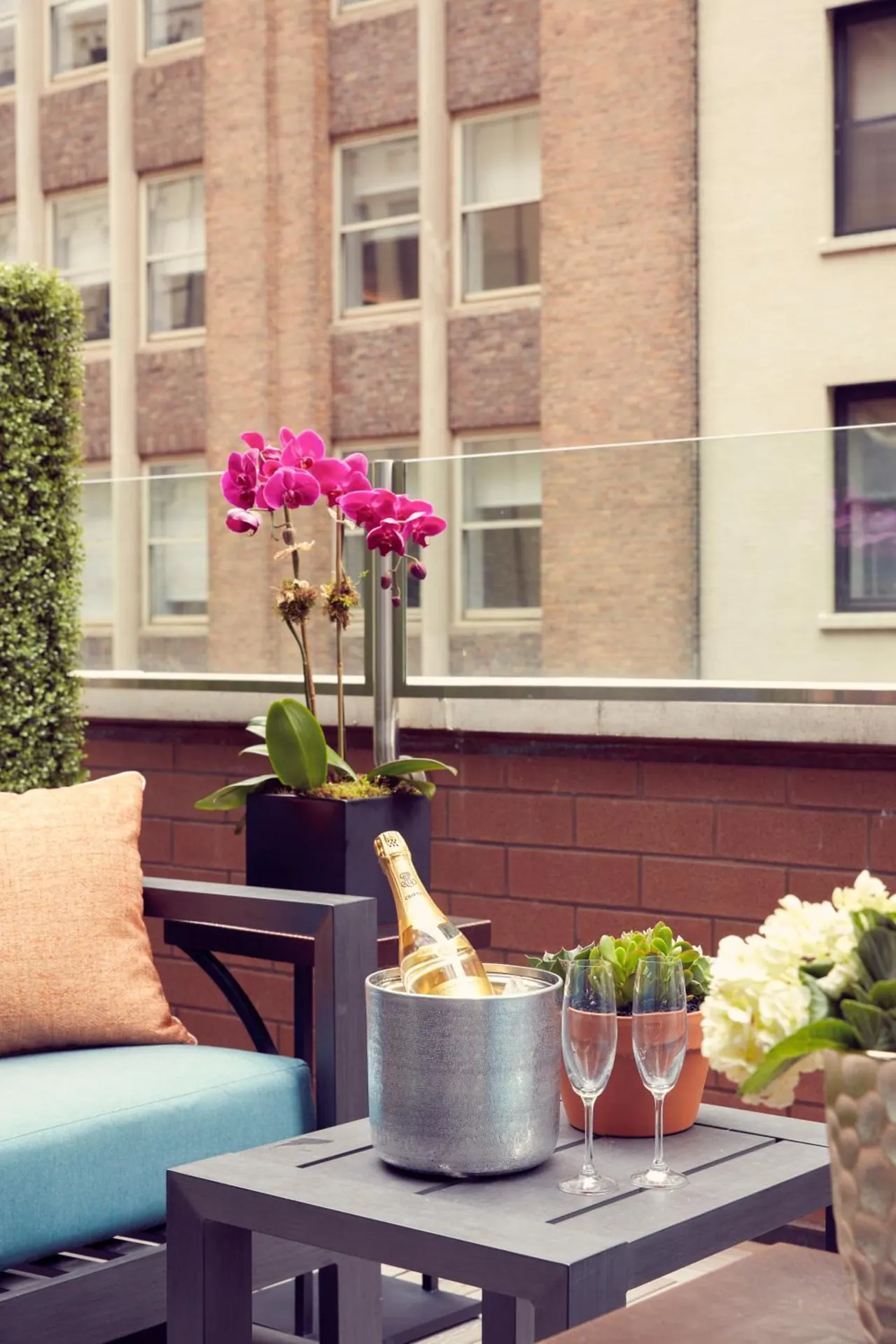 A rooftop terrace with a teal sofa, a side table holding a champagne bottle in an ice bucket and two glasses, pink orchids, and brick buildings beyond the glass railing.