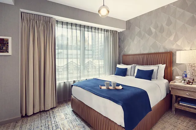 A made bed with blue accents sits beside a window with sheer curtains, with a wooden nightstand and lamp on the right.