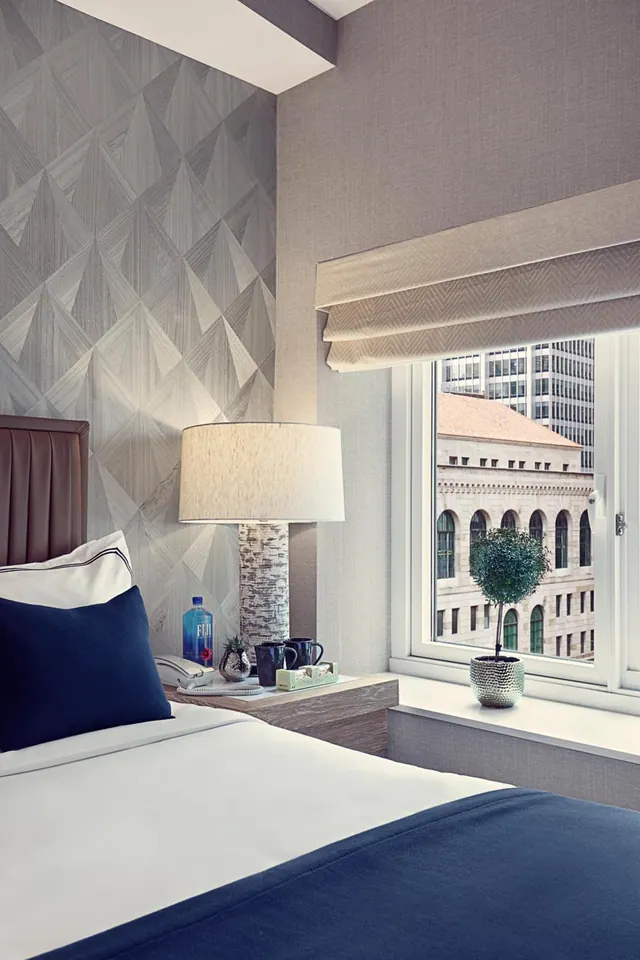 A made bed with navy bedding and pillows sits beside a nightstand with a table lamp, and a window looks out over a stone building with arched windows and a modern tower.