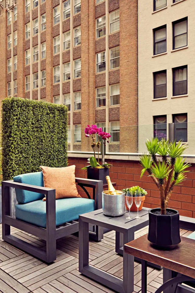 A styled terrace with a blue armchair, orange cushion, potted plants, a champagne bucket and two glasses, with brick and stone apartment buildings behind.