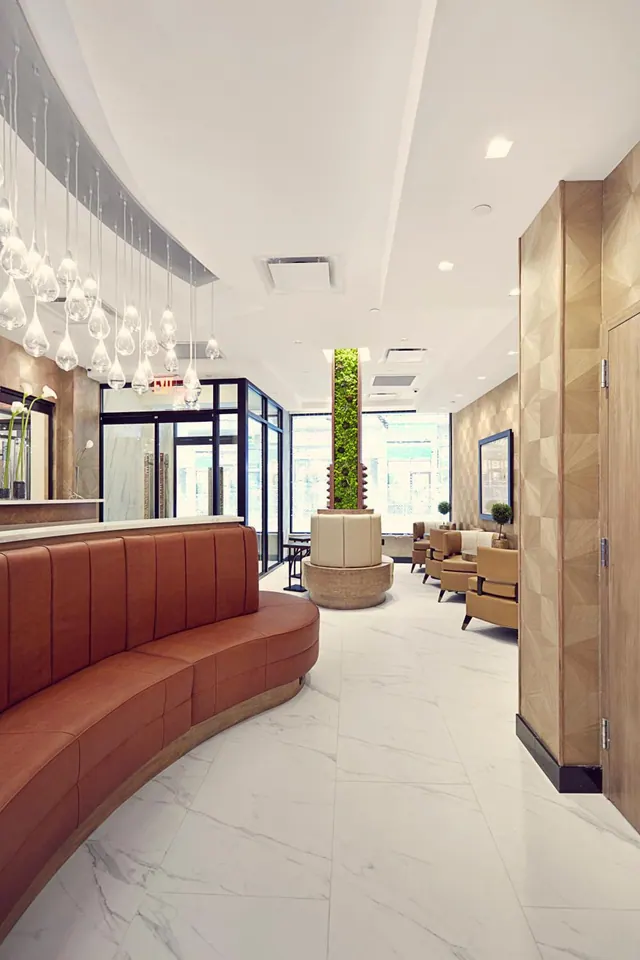A lobby with a curved tan leather banquette, cluster of glass pendant lights, marble floor, a green living wall column, and armchairs near tall windows.