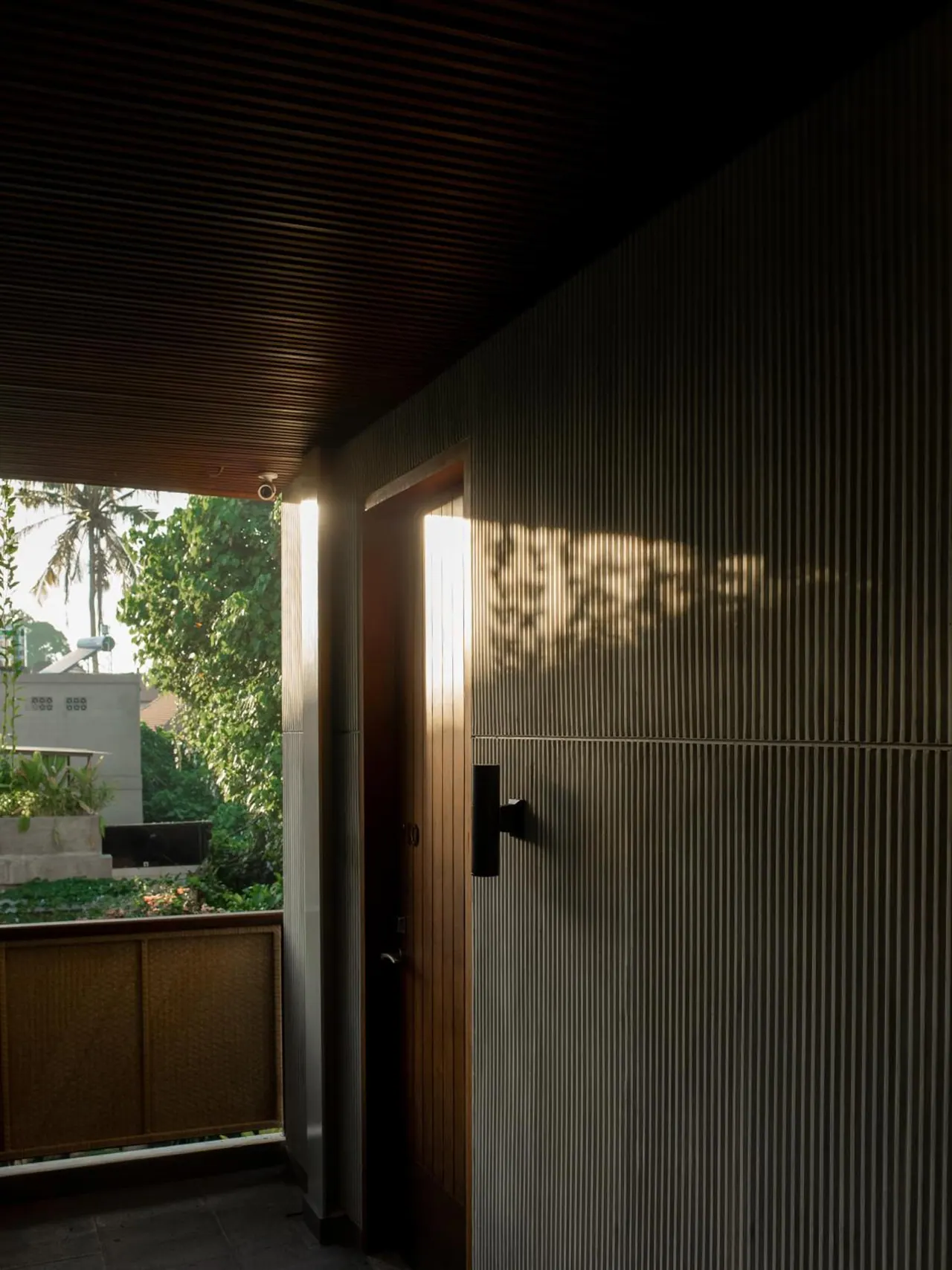 A covered walkway with a ribbed wall and a wooden door, with a view out to garden trees, a palm tree and a concrete wall.