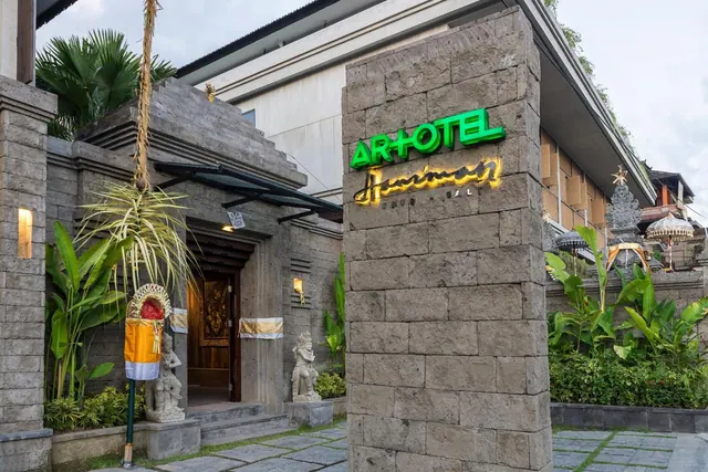 The hotel entrance has a stone pillar with a lit green and gold ART+OTEL Hanoman Bali sign, a stone doorway with a carved statue, and a yellow ceremonial offering beside the steps.