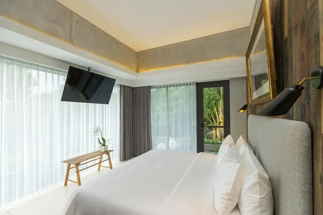 A bedroom with a made king bed, a wall-mounted television, sheer floor-to-ceiling curtains and a glass door opening onto palm trees.