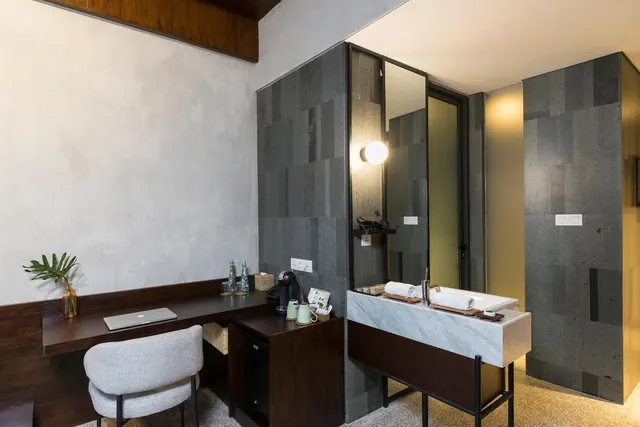 A hotel room with a dark wooden desk, grey upholstered chair, laptop, coffee machine and mini fridge beside a marble vanity with folded towels.