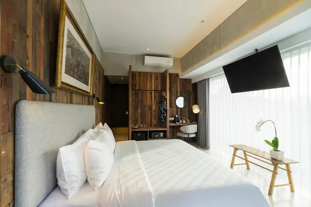 A bedroom with a made bed, wood-panelled walls, a wall-mounted television, a desk with a round mirror and sheer curtains covering floor-to-ceiling windows.