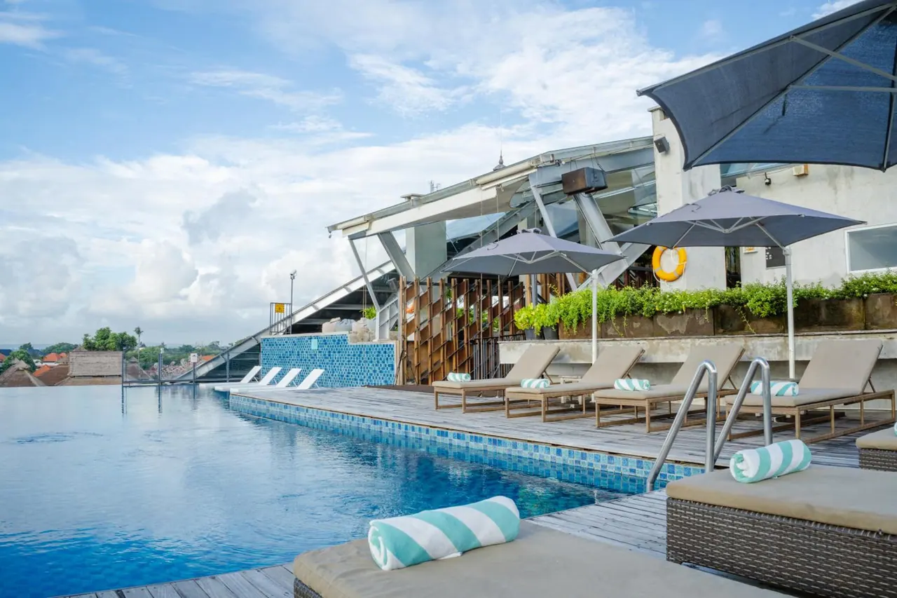 An infinity-edge pool with striped towels on sun loungers and grey umbrellas beside a glass-roofed building, with rooftops and trees in the distance.