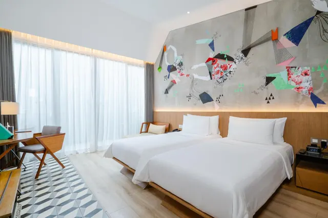 A hotel bedroom with two single beds, a colourful painted wall, a desk with a chair and sheer-curtained windows.