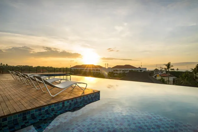 An infinity pool with blue mosaic tiles and white sun loungers on a wooden deck, overlooking rooftops as the sun sets.