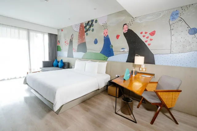 A hotel bedroom with a king bed with white linen, a grey sofa with blue cushions, a painted mural on the wall, and a wooden desk with a chair.