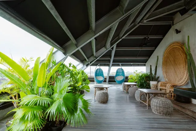 A covered rooftop terrace with wooden decking, two hanging blue egg chairs, rattan seating, woven stools and potted tropical plants.