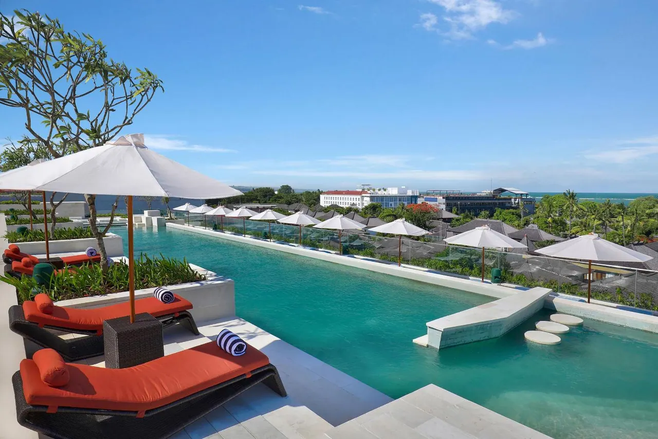 A long rooftop swimming pool with white parasol umbrellas and orange sun loungers, with a view over trees and buildings toward the sea.