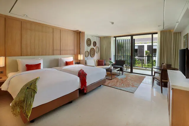 A twin-bedded hotel room with a seating area, a work desk, a television and glass doors opening onto a balcony with greenery beyond.
