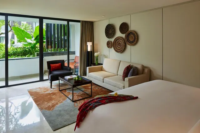 A hotel bedroom with a large bed, a sofa and armchair, and glass doors opening onto a balcony with tropical garden plants.