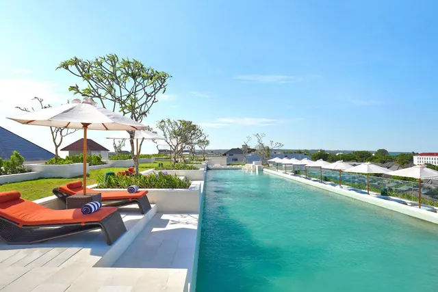 A long swimming pool with white parasols, orange-cushioned sun loungers and frangipani trees on the surrounding deck and lawn.