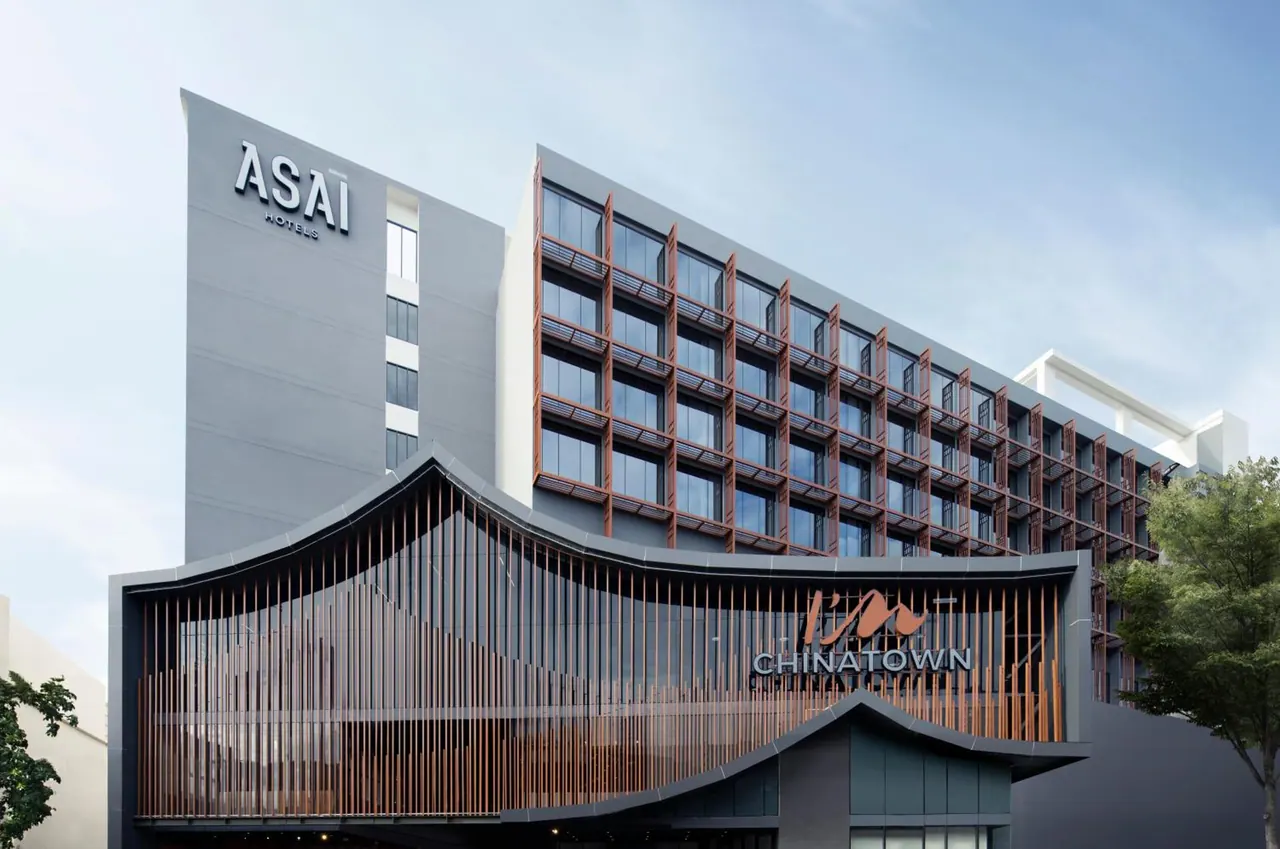 The exterior of the ASAI Hotel Chinatown building shows a curved, wood-slatted entrance canopy below a multi-storey facade of windows under a pale sky.