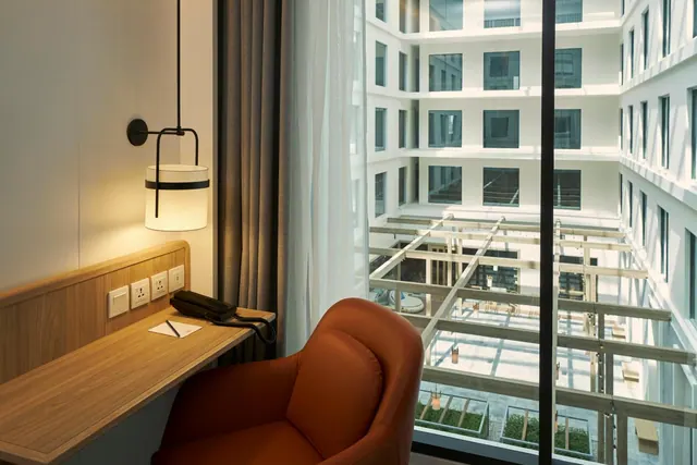 A guest room corner with a wooden desk, a pendant lamp, an orange armchair and a floor-to-ceiling window looking onto an inner courtyard of the building.