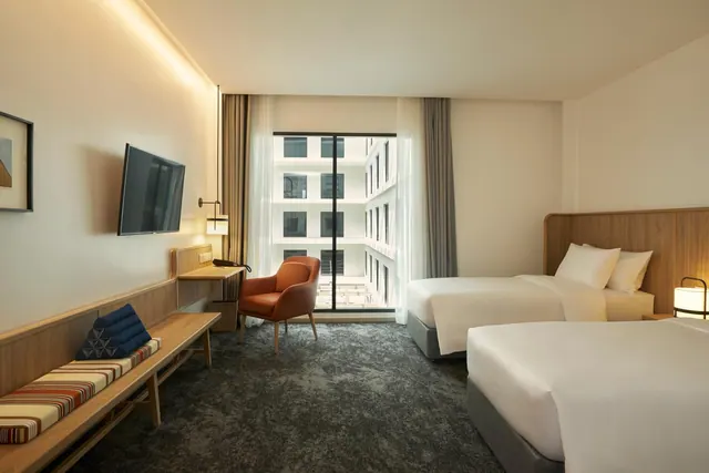 A hotel bedroom with two beds, a wall-mounted TV, a desk, an orange armchair and a floor-to-ceiling window facing another building.