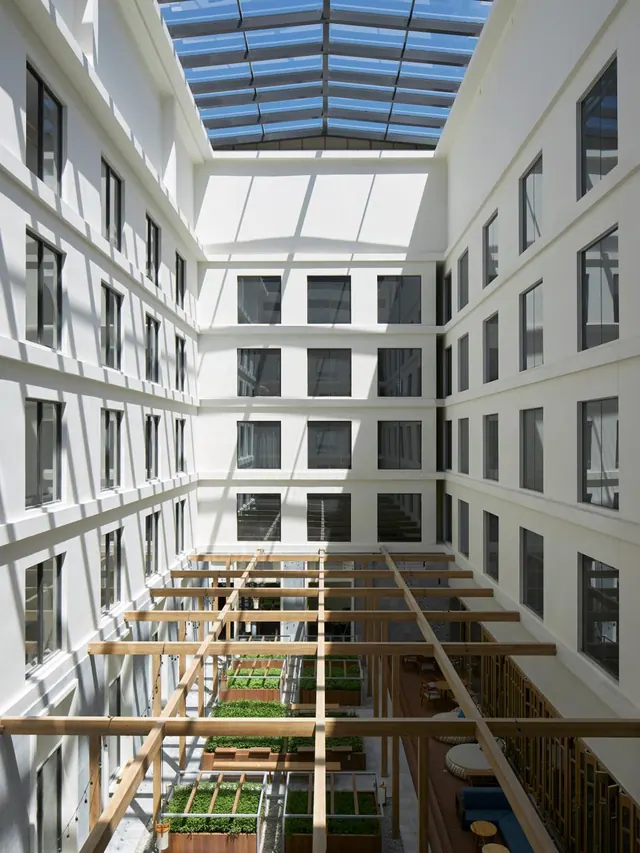 A white multi-storey atrium courtyard with a glass skylight, wooden pergola beams, planted boxes and a seating area with blue cushions at right.