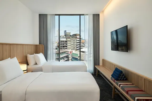 A hotel room with two white beds facing floor-to-ceiling windows that look out over low-rise city buildings, with a wall-mounted TV and a wooden bench with Thai cushions.