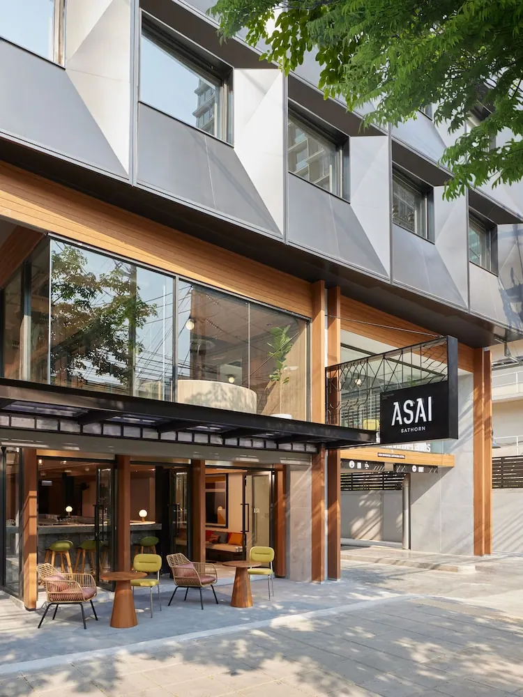 Exterior of the ASAI Sathorn hotel showing a glass-fronted ground floor, outdoor tables and chairs on the forecourt, and a black sign on the facade.