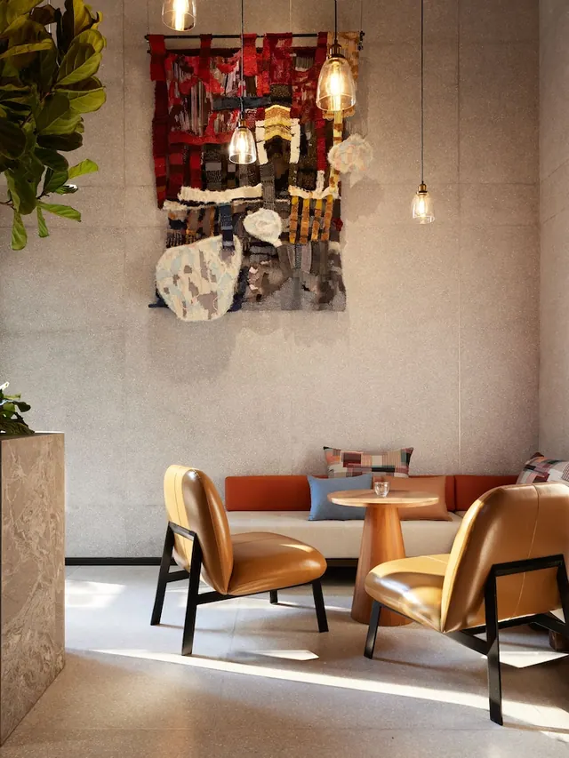 A lounge seating area with two tan leather armchairs, a round wooden side table, an orange-backed sofa and a woven wall hanging under pendant lights.